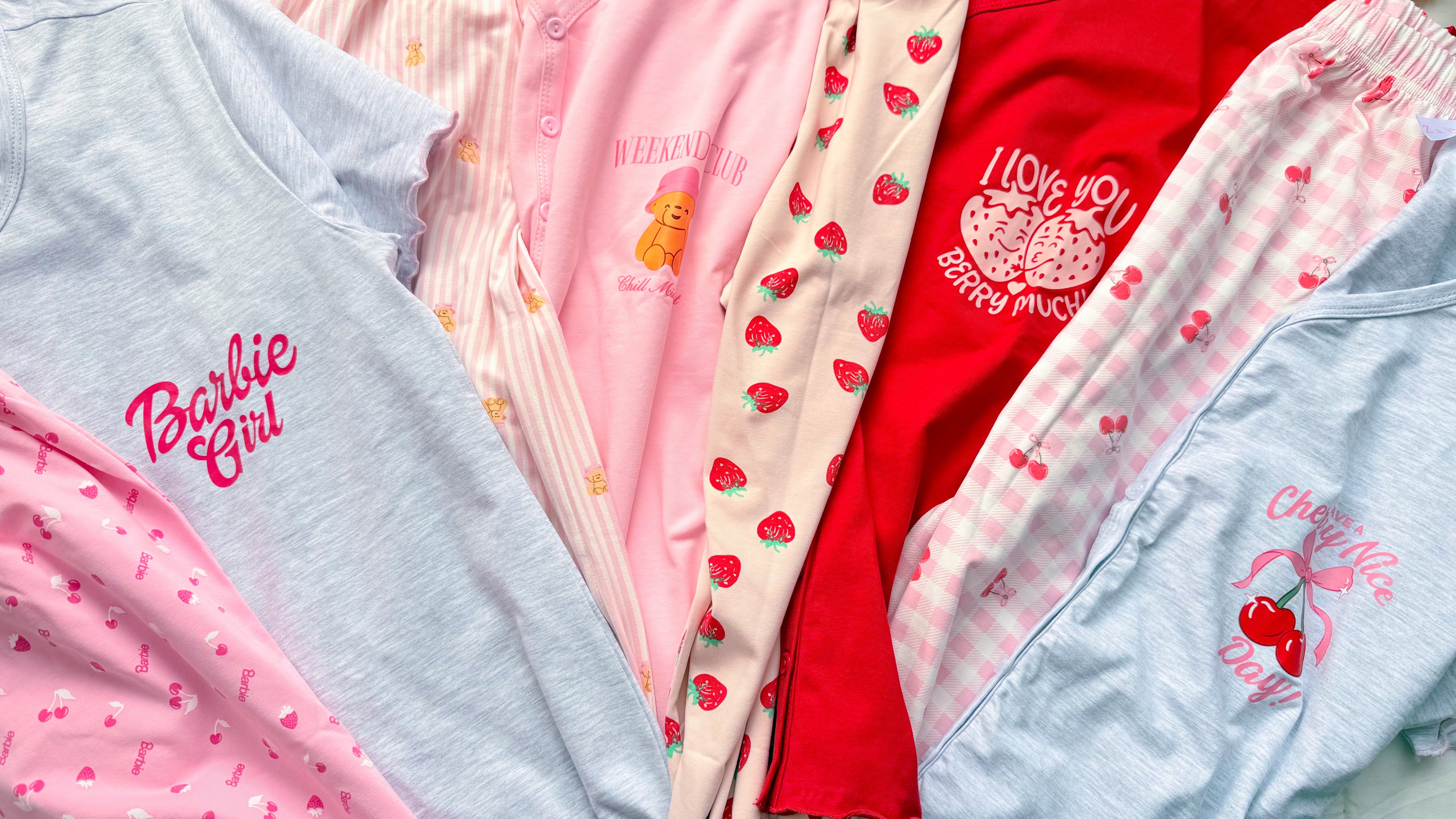 A close-up flat lay of several Barbie-themed pajama tops or sleepwear pieces in pastel and bright colors, including light blue, pink, cream, and red. Visible printed branding includes the Barbie logo and 'Barbie Music' text.